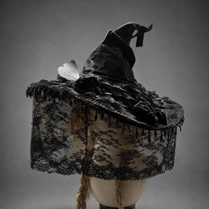 Könnte beinhalten: Ein schwarzer Spitzhut mit breiter Krempe, verziert mit schwarzer Spitzenborte und hängenden Perlen. Der Hut ist seitlich mit einem weißen Schmetterling und oben mit einer schwarzen Schleife versehen. Ein Spitzenschleier hängt vom Rand herab.