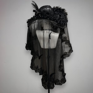 Puede incluir: Un sombrero negro con ala ancha adornado con detalles florales oscuros, plumas y flecos de cuentas colgantes. Un velo negro largo y transparente con bordes de encaje intrincados cuelga del ala, cubriendo un soporte de exhibici&oacute;n de torso de maniqu&iacute;.