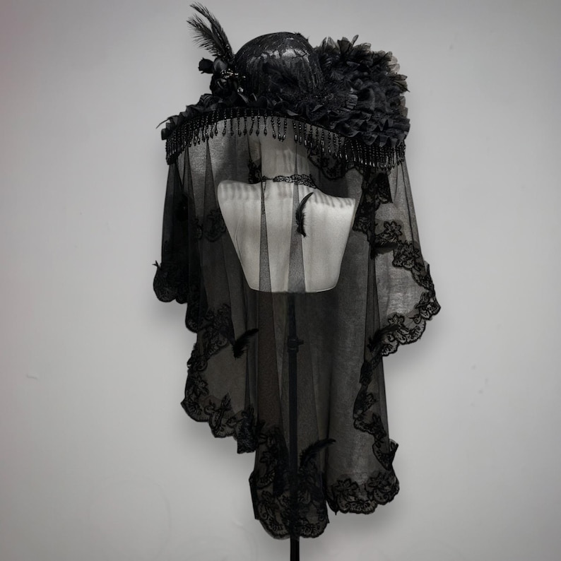 Puede incluir: Un sombrero negro con ala ancha adornado con detalles florales oscuros, plumas y flecos de cuentas colgantes. Un velo negro largo y transparente con bordes de encaje intrincados cuelga del ala, cubriendo un soporte de exhibici&oacute;n de torso de maniqu&iacute;.