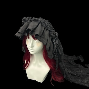 May include: A black headpiece featuring a wide, ruffled fabric brim and decorative fabric roses on the sides. The headpiece includes a long, flowing lace veil that extends from the back, displayed on a mannequin with long red hair.