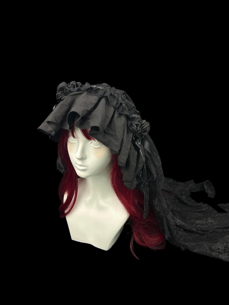 May include: A black headpiece featuring a wide, ruffled fabric brim and decorative fabric roses on the sides. The headpiece includes a long, flowing lace veil that extends from the back, displayed on a mannequin with long red hair.