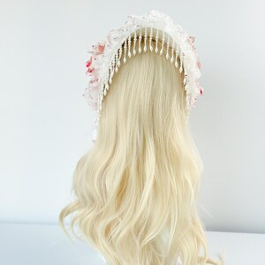 May include: A white bridal headpiece with delicate lace, pink floral accents, and dangling beaded strands. The headpiece is displayed on a blonde wig, showcasing its design and details. The headpiece is a bridal accessory.