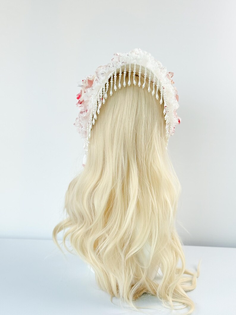 May include: A white bridal headpiece with delicate lace, pink floral accents, and dangling beaded strands. The headpiece is displayed on a blonde wig, showcasing its design and details. The headpiece is a bridal accessory.