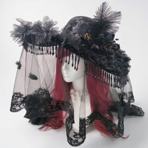 Puede incluir: Un sombrero negro de ala ancha con detalles de encaje intrincados, adornos florales oscuros y plumas erguidas. El sombrero est&aacute; decorado con flecos de cuentas negras colgantes y un velo negro largo y transparente con borde de encaje.