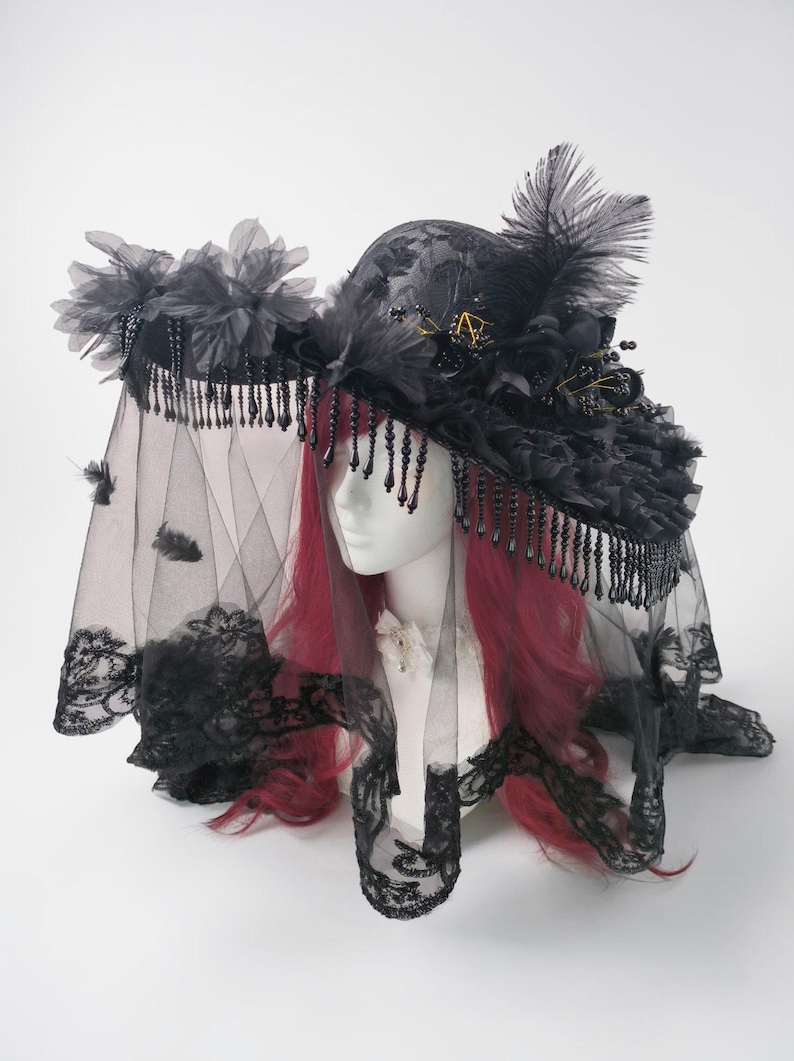 Puede incluir: Un sombrero negro de ala ancha con detalles de encaje intrincados, adornos florales oscuros y plumas erguidas. El sombrero est&aacute; decorado con flecos de cuentas negras colgantes y un velo negro largo y transparente con borde de encaje.