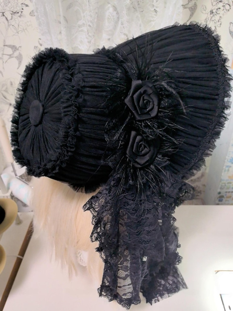 Op de afbeelding: Een zwarte bonnet met een geplooid ontwerp, versierd met twee stoffen rozen en veren. De bonnet heeft een kanten rand en een kanten draperie. De hoed wordt getoond op een mannequinhoofd.