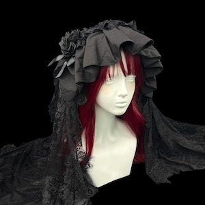 May include: A black headpiece featuring a ruffled fabric crown adorned with fabric roses and long, intricate black lace veils that drape down the sides. The headpiece is displayed on a mannequin head with red hair against a black background.