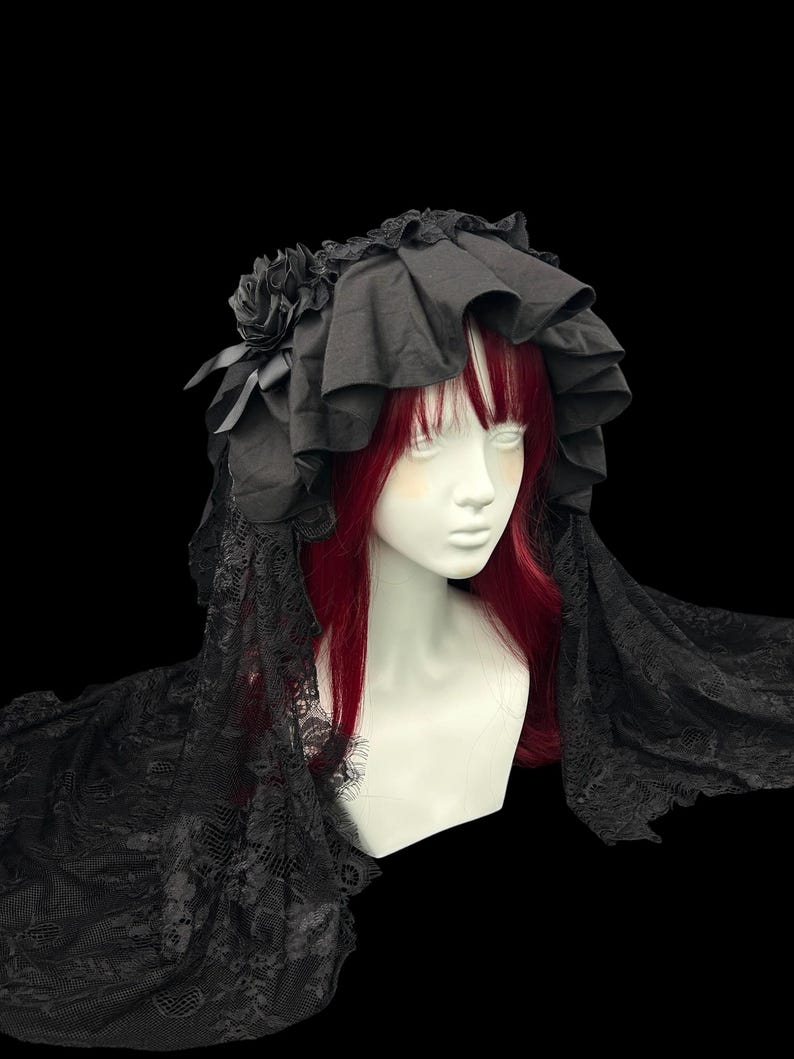 May include: A black headpiece featuring a ruffled fabric crown adorned with fabric roses and long, intricate black lace veils that drape down the sides. The headpiece is displayed on a mannequin head with red hair against a black background.