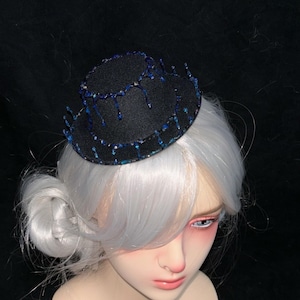 May include: A small, black hat with a beaded, dripping blue design. The hat is worn on a mannequin head with white hair styled in a bun. The mannequin has pale skin and pink makeup.