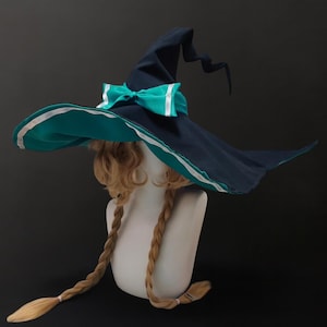 May include: A dark blue witch hat with a wide brim, teal underside, and a white stripe along the edge. A teal bow with white accents is at the front. The hat is displayed on a mannequin head with blonde braids, suitable for a costume or cosplay.