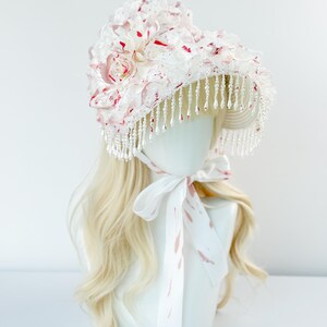 May include: A white bonnet with red accents and a beaded fringe. The bonnet is adorned with fabric flowers and a matching ribbon tie. The bonnet is displayed on a mannequin head with blonde hair.
