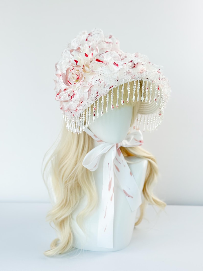 May include: A white bonnet with red accents and a beaded fringe. The bonnet is adorned with fabric flowers and a matching ribbon tie. The bonnet is displayed on a mannequin head with blonde hair.