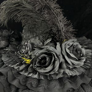 Puede incluir: Un sombrero negro con una copa alta y puntiaguda adornada con una gran pluma de avestruz negra erguida. El ala est&aacute; decorada con volantes de tela negra superpuestos, encaje y dos grandes rosas de tela negra con peque&ntilde;as cuentas negras.