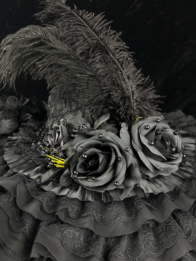 Puede incluir: Un sombrero negro con una copa alta y puntiaguda adornada con una gran pluma de avestruz negra erguida. El ala est&aacute; decorada con volantes de tela negra superpuestos, encaje y dos grandes rosas de tela negra con peque&ntilde;as cuentas negras.
