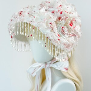 May include: A white bonnet with red floral accents and pearl bead fringe. The bonnet features layers of ruffled fabric and a matching ribbon tie. The design is a mix of textures and colors, creating a unique and elegant look.