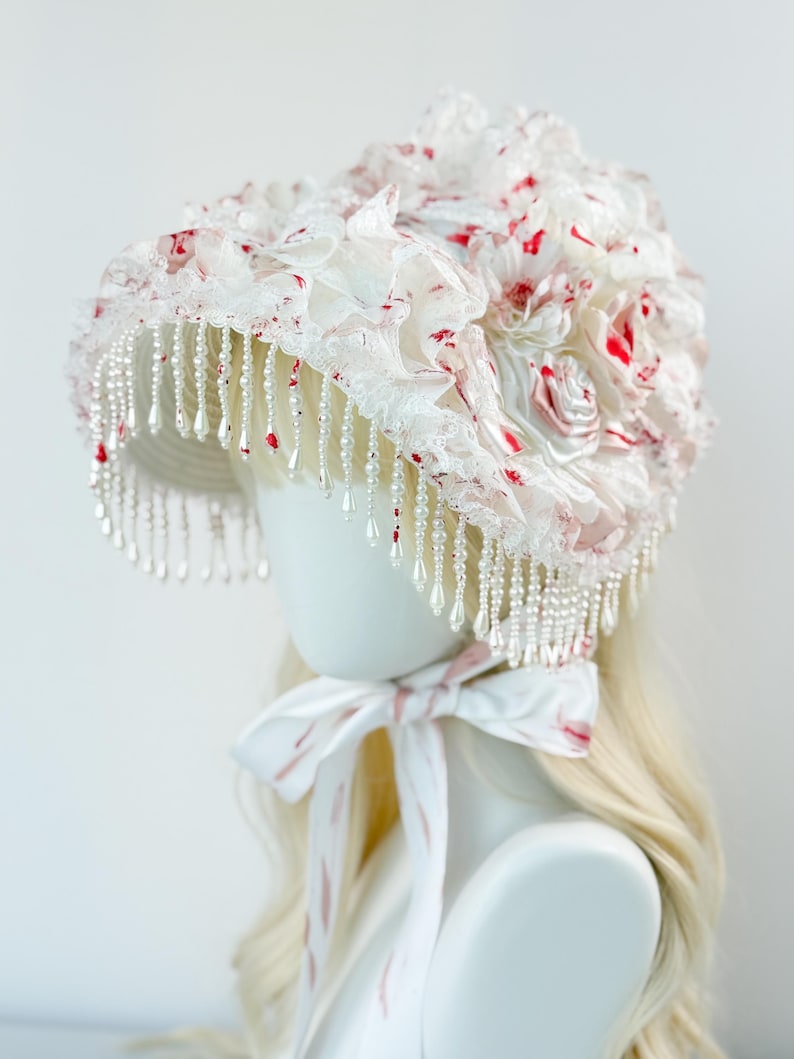 May include: A white bonnet with red floral accents and pearl bead fringe. The bonnet features layers of ruffled fabric and a matching ribbon tie. The design is a mix of textures and colors, creating a unique and elegant look.