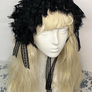 May include: A black lace bonnet featuring intricate floral patterns and scalloped edges. The headpiece is constructed with layered, ruffled lace fabric and includes two long, narrow lace ribbons that hang down from the sides to tie under the chin.