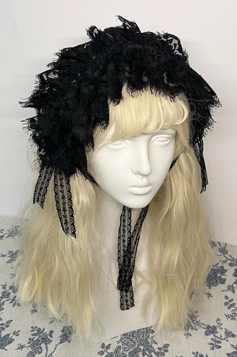 May include: A black lace bonnet featuring intricate floral patterns and scalloped edges. The headpiece is constructed with layered, ruffled lace fabric and includes two long, narrow lace ribbons that hang down from the sides to tie under the chin.