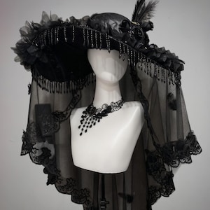 Puede incluir: Un sombrero negro de ala ancha adornado con rosas de tela, plumas y cuentas negras colgantes en el borde. Un velo negro transparente con un intrincado borde de encaje cuelga del ala, junto con una gargantilla de encaje negro a juego.