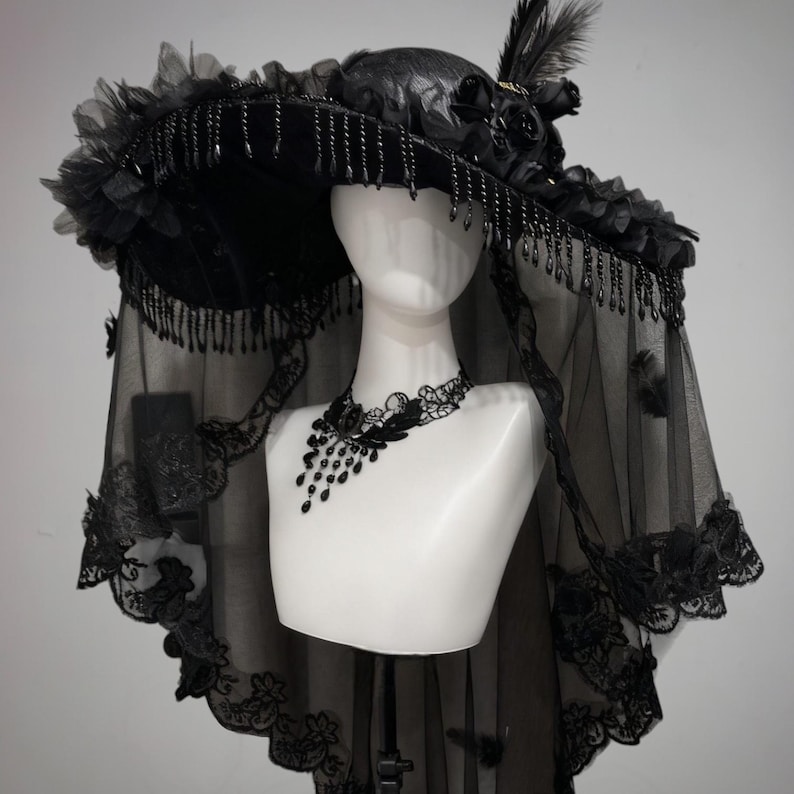 Puede incluir: Un sombrero negro de ala ancha adornado con rosas de tela, plumas y cuentas negras colgantes en el borde. Un velo negro transparente con un intrincado borde de encaje cuelga del ala, junto con una gargantilla de encaje negro a juego.