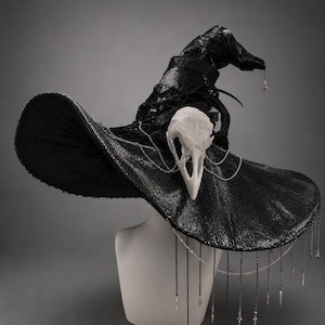 Peut inclure: Un chapeau de sorcière noir texturé orné d'un crâne d'oiseau blanc sur le devant. Le chapeau est décoré de chaînes argentées, de dentelle noire et de breloques en forme de croix argentées pendantes sur le large bord et la pointe.