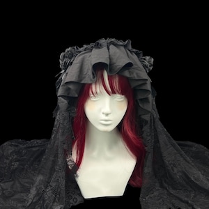May include: A black headpiece featuring a ruffled fabric crown adorned with side rosettes and a long, intricate lace veil that drapes over the shoulders. The headpiece is displayed on a mannequin head with straight, vibrant red hair and bangs.