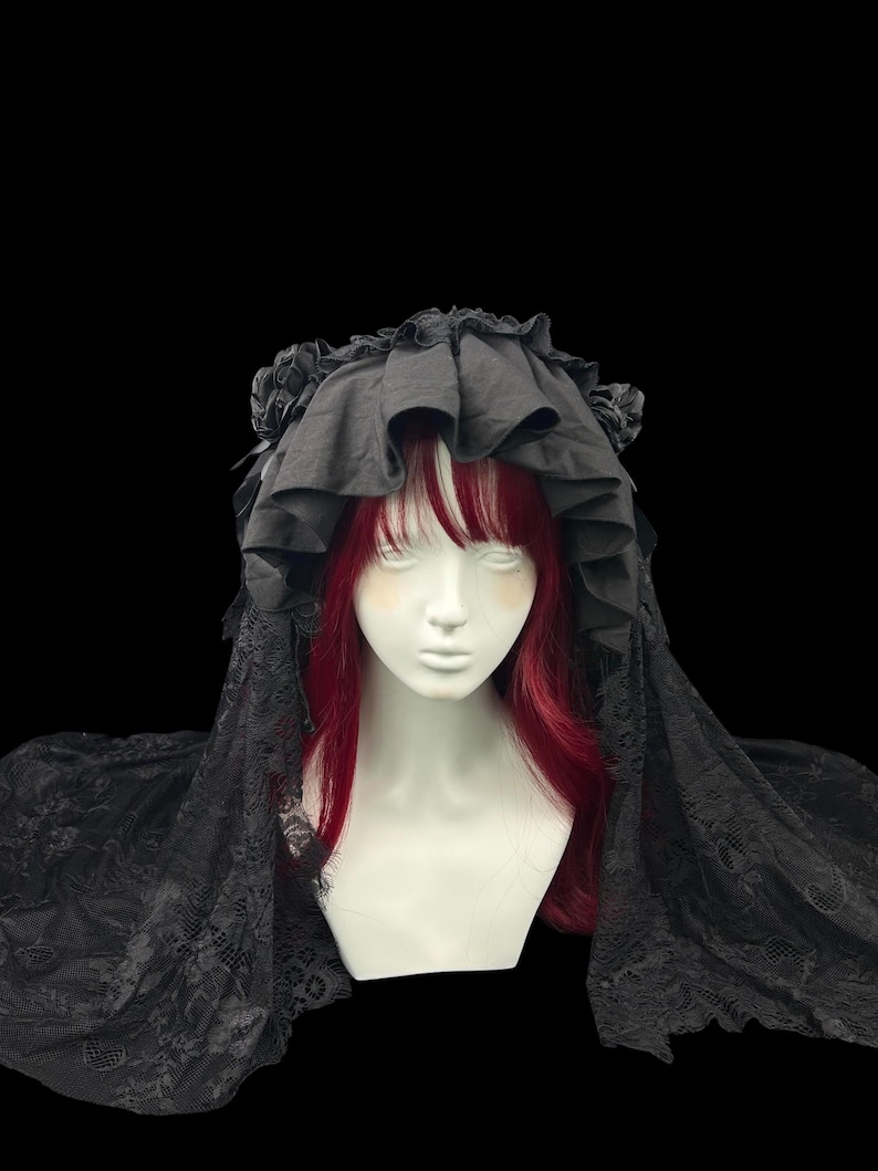 May include: A black headpiece featuring a ruffled fabric crown adorned with side rosettes and a long, intricate lace veil that drapes over the shoulders. The headpiece is displayed on a mannequin head with straight, vibrant red hair and bangs.