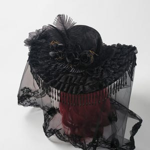 Puede incluir: Un sombrero negro con un ala ancha y con volantes adornado con rosas de tela negra, plumas y flecos de cuentas colgantes. Un largo velo de encaje negro transparente con intrincados bordados florales cuelga del ala sobre una cabeza de maniqu&iacute; pelirroja.