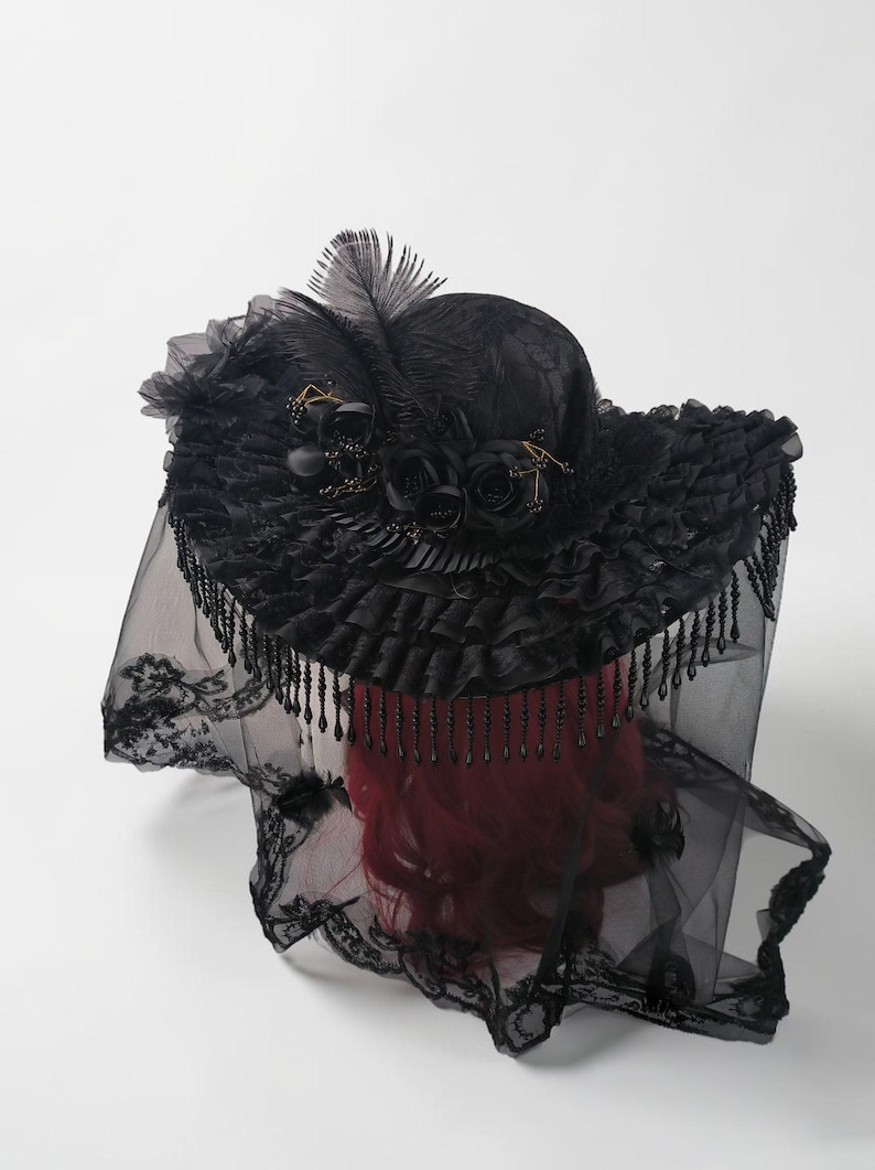 Puede incluir: Un sombrero negro con un ala ancha y con volantes adornado con rosas de tela negra, plumas y flecos de cuentas colgantes. Un largo velo de encaje negro transparente con intrincados bordados florales cuelga del ala sobre una cabeza de maniqu&iacute; pelirroja.