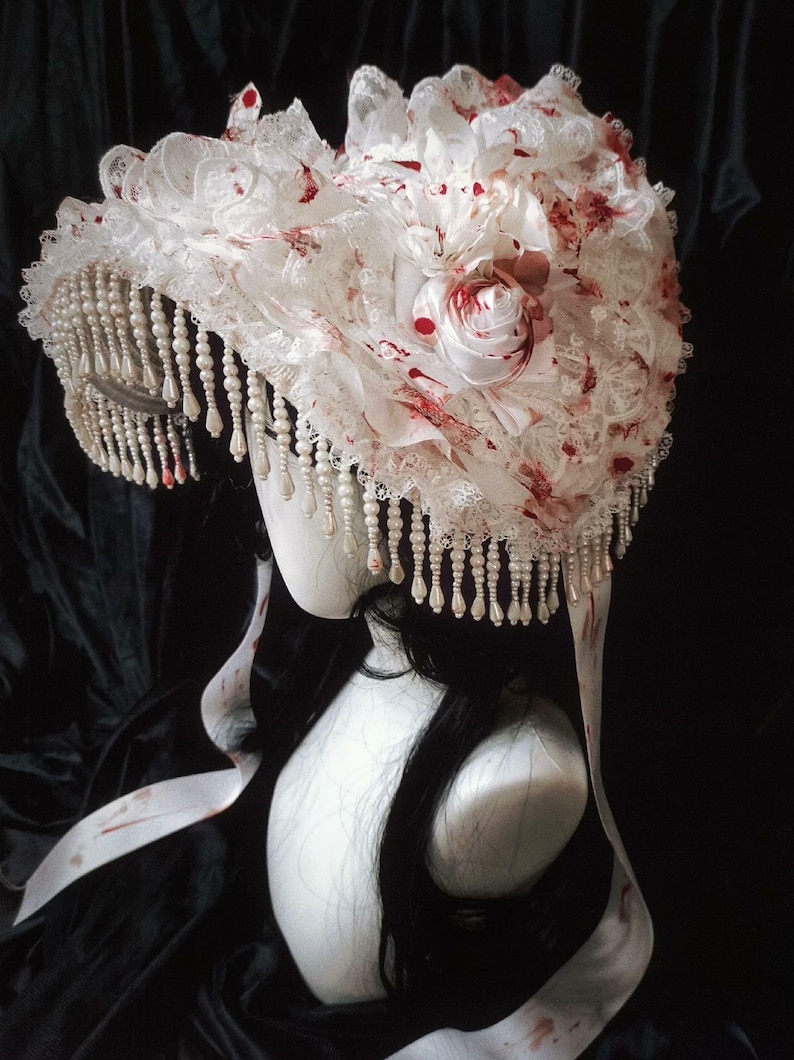 May include: A white headpiece with lace, fabric flowers, and faux pearls. The headpiece has red accents and a beaded fringe. The design is on a mannequin head with long black hair and white ribbons.