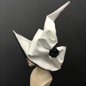 May include: A tall, pointed white hat with a wide, curved brim and a large, structured white bow at the front. A single black rose is attached to the centre of the bow. The hat features a black trim along the edge of the brim and a smooth finish.