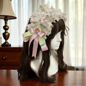 May include: A white and gold lace headpiece with a ruffled design. The headpiece features a pink and green bow with a pink jewel in the center. The headpiece is displayed on a white mannequin head with brown hair.