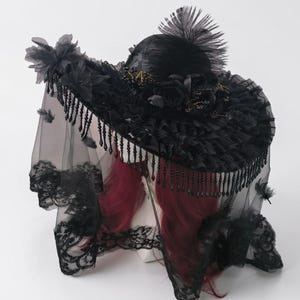 Puede incluir: Un sombrero negro de ala ancha con una copa alta adornada con una gran pluma, rosas de tela y detalles dorados. El ala est&aacute; decorada con tela fruncida y cuentas negras colgantes, complementado por un velo de encaje transparente con flores.