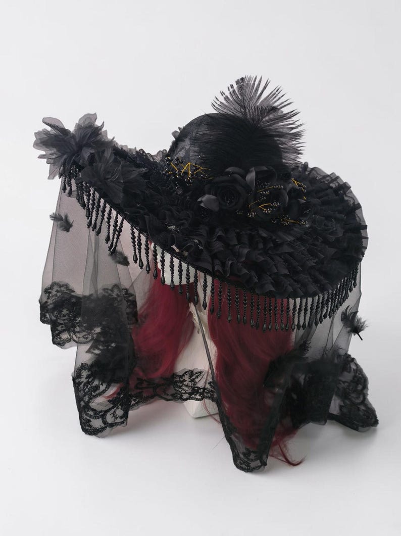 Puede incluir: Un sombrero negro de ala ancha con una copa alta adornada con una gran pluma, rosas de tela y detalles dorados. El ala est&aacute; decorada con tela fruncida y cuentas negras colgantes, complementado por un velo de encaje transparente con flores.