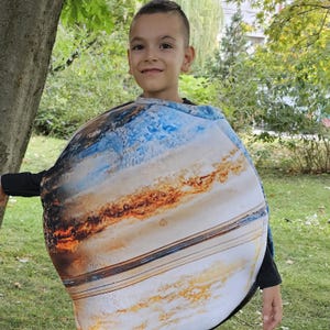 Può includere: Un bambino indossa un costume del pianeta Giove. Il costume è un design rotondo e imbottito con una stampa dettagliata delle fasce nuvolose vorticose del pianeta in tonalità di blu, marrone e bianco. Il bambino sorride su uno sfondo verde.