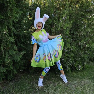 Puede incluir: Una persona con un disfraz de Pascua colorido, que incluye un sombrero de orejas de conejo blanco, una camiseta con un diseño de conejo, una falda y leggings. El atuendo presenta colores pastel y diseños de huevos de Pascua. Zapatillas blancas completan el look.