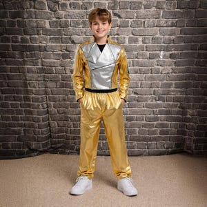 Op de afbeelding: Een goudkleurig kostuum met een zilver-gouden jasje en bijpassende broek. De jas heeft gouden knopen en een zwarte bies. De persoon draagt witte sneakers. De achtergrond is een bakstenen muur.