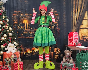 Puede incluir: Una persona con un disfraz de elfo verde con un gorro rojo y verde, una túnica verde y una falda verde. El disfraz incluye un cinturón negro, mallas verdes y zapatos de elfo verdes. El fondo presenta un árbol de Navidad, regalos envueltos y una chimenea.