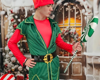 Puede incluir: Un disfraz de elfo navideño con una túnica verde con ribetes dorados, una camisa roja de manga larga y un cinturón negro con hebilla dorada. El elfo lleva un gorro rojo y verde y sostiene un accesorio a rayas verdes y blancas.