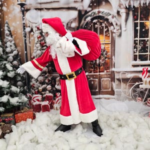 Può includere: Babbo Natale in un abito rosso con finiture bianche, una cintura nera con fibbia dorata e un cappello rosso. Porta una grande sacca rossa sulla spalla. Lo sfondo presenta una scena innevata con decorazioni natalizie.