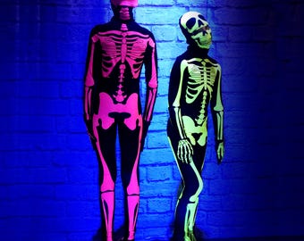 May include: Two full-body skeleton costumes. One is black with neon pink skeleton detailing, including a head covering. The other is black with neon green skeleton detailing, also with a head covering. Both costumes are standing in front of a blue brick wall.
