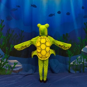 Peut inclure: Un costume de tortue pour enfant, composé d'une combinaison verte, d'une carapace jaune et verte et d'un couvre-chef de tortue. Le costume a des manches en forme de nageoires. Le fond est une scène sous-marine bleue avec des poissons.