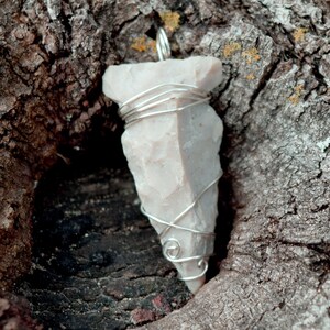 May include: A white stone arrowhead pendant wrapped in silver wire. The pendant is hanging from a tree branch.