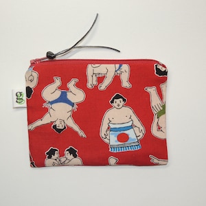May include: Red zippered pouch with a repeating pattern of cartoon sumo wrestlers in various poses. The pouch has a black zipper pull.