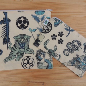 May include: Two beige zippered pouches with a Japanese-inspired design. The larger pouch features a tiger, a snake, a flower, and a wave. The smaller pouch features a scorpion, a snake, a flower, and a woman.