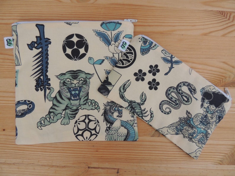 May include: Two beige zippered pouches with a Japanese-inspired design. The larger pouch features a tiger, a snake, a flower, and a wave. The smaller pouch features a scorpion, a snake, a flower, and a woman.