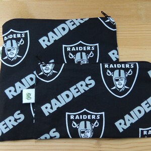 May include: Two black zippered pouches with a white and gray repeating pattern of the Las Vegas Raiders logo. The logo features a football player's head with a pirate eye patch and crossed swords.