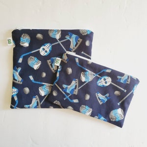May include: Two reusable zippered pouches with a hockey theme. The pouches are navy blue with a white zipper and a white background for the hockey stick, puck, and skate designs. The pouches are made of fabric and have a white background with the text "GD" in green.
