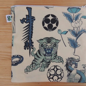 May include: A cream-colored zippered pouch with a Japanese-inspired design. The pouch features a tiger, a dragon, a snake, flowers, and geometric shapes in shades of blue and black.