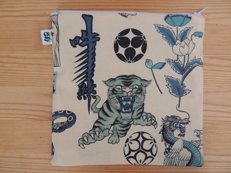 May include: A cream-colored zippered pouch with a Japanese-inspired design. The pouch features a tiger, a dragon, a snake, flowers, and geometric shapes in shades of blue and black.